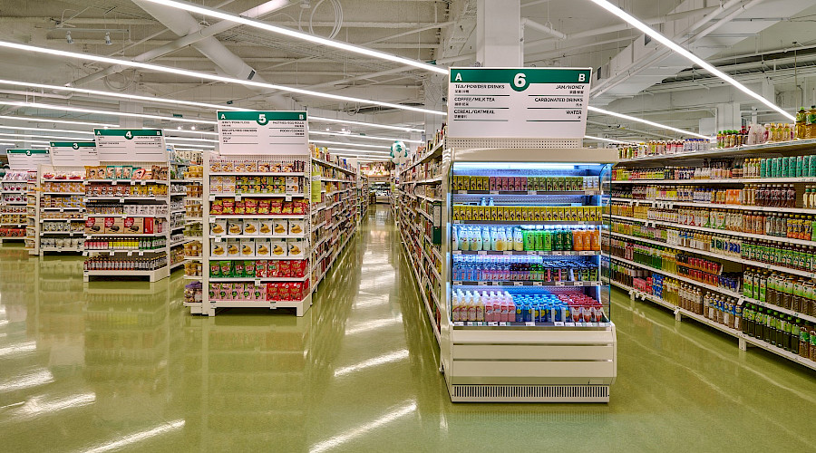 Rows of shelves of products in a supermarket with bright lights and green flooring.