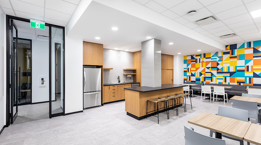 Interior shot of the staff break room and kitchen at the Canadian Western Bank in Lethbridge, Alberta, built by Chandos Construction Calgary. The accent wall is decorated with bright shades of orange, blue, and white with modern shapes.